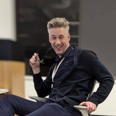 Michael Hawks, sentado, suéter navy, sonriendo con gafas en la mano